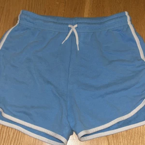 Blå shorts med vit kant Cubus  - Snygga ljusblå shorts med vita kanter och snörning i midjan. Perfekta för träning eller varma dagar. Mjukt material och klassisk sportig look.