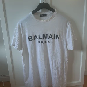 Vit t-shirt från Balmain  - Säljer en klassisk vit t-shirt från Balmain med svart tryck framtill. T-shirten har rund halsringning och korta ärmar. Perfekt för en stilren och trendig look.