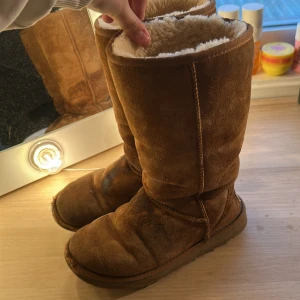 UGG boots - Säljer ett par bruna UGG-boots  i mocka med mjukt vitt fårskinnsfoder. Skorna har platt sula och hög skaft, perfekta för kalla dagar. UGG-logga bak på hälen.