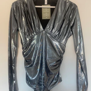 Glittrig silver body från H&M - Säljer en glittrig silverfärgad body från H&M med djup v-ringning och långa ärmar. Toppen har draperad effekt framtill och är tillverkad i återvunnen polyester med elastan. Perfekt för att sticka ut på festen!