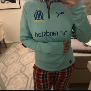 Ljusblå Marseille träningströja från Puma - Säljer en ljusblå långärmad träningströja från Puma med Olympique de Marseille-logga och Randstad-tryck på bröstet. Tröjan har halv dragkedja och sportig design, perfekt för fotbollsträning.