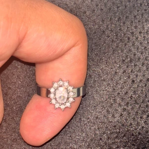 Silverfärgad ring med glittrande stenar - Säljer en elegant silverfärgad ring med en oval, klar sten i mitten omgiven av små gnistrande stenar i en blomliknande form. Perfekt för dig som vill ha något stilrent och glittrigt.