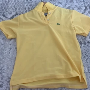 Gul piké från Lacoste - Snygg gul pikétröja från Lacoste med klassisk krage och två knappar framtill. Ikoniska gröna krokodilloggan broderad på bröstet. Perfekt för en fräsch och sportig look.