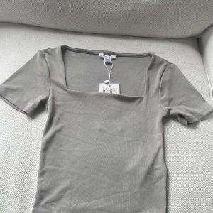 Ljusgrå topp med fyrkantig ringning  - Säljer en ljusgrå topp med korta ärmar och snygg fyrkantig ringning. Toppen har en tight passform och är tillverkad i ett mjukt, stretchigt material. Perfekt att styla till jeans eller kjol.  Etikett finns kvar på plagget