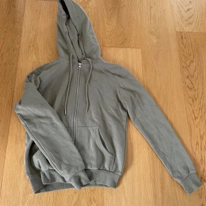 Grön  hoodie med dragkedja - Säljer en grön hoodie med dragkedja och huva. Den har långa ärmar, ribbade muddar och fickor framtill. Perfekt för en avslappnad stil.