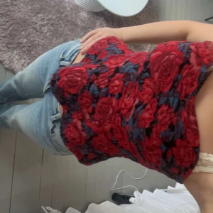 Röd blommig bandue topp - Säljer en röd bandue topp med blommigt mönster och svarta detaljer. Toppen är ärmlös och har en figurnära passform, perfekt att styla med jeans eller kjol. Supersnygg och trendig detalj i garderoben!