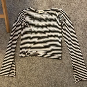 Randig långärmad topp från Gina Tricot - Säljer en svartvit randig långärmad topp från Gina Tricot. Tröjan har smala horisontella ränder och slitsar med knappdetaljer vid ärmsluten. Perfekt för en stilren och enkel look.
