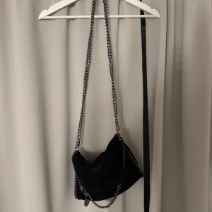 Svart axelväska med kedjerem - Säljer en svart axelväska med snygg kedjerem och mjukt material. Väskan har en cool design och passar perfekt för dig som vill ha en stilren accessoar med lite edge. Perfekt storlek för det viktigaste!
