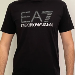 Svart t-shirt från Emporio Armani EA7 - Snygg svart t-shirt från Emporio Armani EA7 med stort logotryck på bröstet. Klassisk passform och rund halsringning. T-shirten är i storlek L men har i passform för S/M! T shirten är ÄKTA. Denna T shirt är en limiterad produktion av EA7.