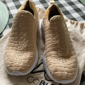 Beige sneakers från MoCobler - Säljer ett par beige sneakers från MoCobler med quiltad struktur och vit sula. Skorna är slip-on och har märkets logga broderad på ovansidan. Perfekta för en avslappnad och trendig stil.