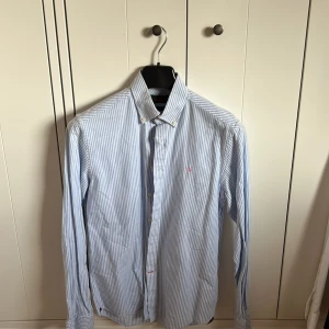 Morris skjorta - Säljer en klassisk blårandig skjorta från Morris i storlek M. Skjortan har lång ärm, knappar framtill och en liten rosa broderad detalj på bröstet. Tillverkad i bomull och passar perfekt till en stilren look.