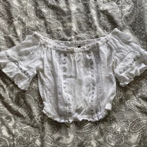 Vit croppad offshoulder blus från H&M Divided - Superfin vit croppad blus från H&M Divided med offshoulder-modell och volangdetaljer på ärmarna. Blusen har spetsdetaljer framtill och är perfekt för sommardagar. Luftigt material och snygg passform.