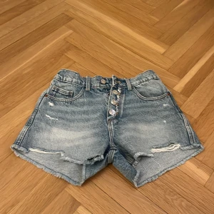 Blå jeansshorts med knappar från Zara - Snygga blå jeansshorts från Zara med hög midja, slitna detaljer och fransig kant. Knappar framtill istället för dragkedja och två bakfickor. Perfekta för sommaren!
