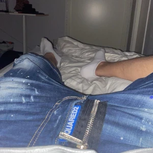 Blå Dsquared2 jeansshorts med färgstänk - Säljer ett par blå jeansshorts från Dsquared2 med coola färgstänk och slitningar. Klassisk femficksmodell med dragkedja och tydlig Dsquared2-logga vid gylfen. Perfekta för en avslappnad och trendig stil.