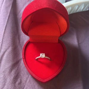 Silverfärgad ring med sten i hjärtformad ask - Säljer en elegant silverfärgad ring med en gnistrande klar sten, levereras i en röd hjärtformad sammetsask. Perfekt som gåva eller promisering. Ringen har en klassisk design med små stenar längs bandet. 