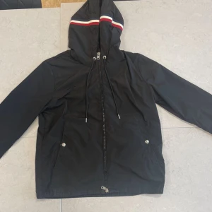 Moncler Wind breaker  - Svart vindjacka från Moncler med huva som har röd, vit och blå rand. Jackan har dragkedja framtill, två fickor med knappar och Moncler-logga på ärmen. Perfekt för blåsiga dagar och snygg streetstil.