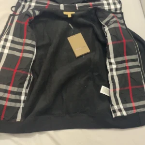 Svart hoodiejacka med rutig huva från Burberry - Säljer en svart hoodiejacka från Burberry med dragkedja och klassiskt rutigt foder i huvan. Jackan har två fickor framtill och ribbade muddar. Perfekt för dig som gillar stilrena detaljer och ikoniskt mönster.