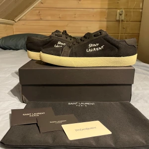 Svarta sneakers från Saint Laurent - Säljer ett par svarta Saint Laurent sneakers med vit sula och broderad logga på sidan. Skorna har snörning och är tillverkade i canvas med detaljer i läder. Klassisk och stilren design med rund tå och platt sula.