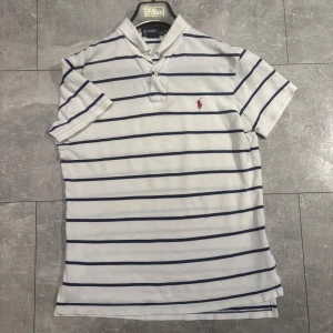 Vit och marinblå randig pikétröja från Polo Ralph Lauren - Vit pikétröja med marinblå ränder från Polo Ralph Lauren. Klassisk krage, korta ärmar och broderad röd logga på bröstet. Perfekt för en stilren och sportig look. dm vid frågor elr fler bilder