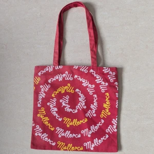 Röd tygkasse med Mallorca-tryck - Säljer en röd tygkasse med spiralformat tryck där det står 'Mallorca' i vitt och gult. Väskan har långa handtag och är perfekt för att bära böcker eller shopping. Enkel och snygg design som sticker ut. Väskan har aldrig använts 