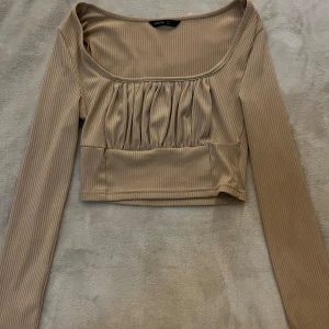 Beige croppad långärmad topp från Shein - Trendig beige croppad topp från Shein med långa ärmar och rundad urringning. Toppen har ribbad struktur och fina rynkade detaljer framtill som ger en snygg passform. Perfekt att styla med höga jeans eller kjol.