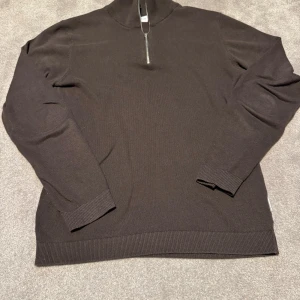 Lindbergh halv zip  - Mörkbrun stickad tröja med halv dragkedja vid halsen. Tröjan har långa ärmar och ribbade muddar vid ärmslut och nederkant. Perfekt för lager-på-lager och enkel att matcha med olika outfits.