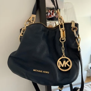 Äkta Marinblå handväska från Michael Kors - Säljer min snygga äkta marinblå handväska från Michael Kors med guldfärgade metalldetaljer och MK-hänge. Väskan är tillverkad i skinn och har dragkedja upptill. Perfekt för dig som vill ha en stilren och klassisk accessoar.