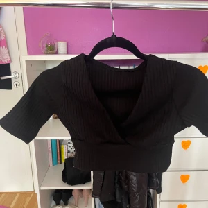 Svart croppad omlottopp - Säljer en svart croppad omlottopp med ribbad struktur. Toppen har korta ärmar och en djup v-ringning framtill. Perfekt att styla med höga jeans eller kjol. Står ingen storlek på plagget men skulle gissa på xxs, möter bara upp i Sthlm och tar Swish eller kontanter🫶🏻