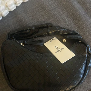 Svart axelväska från beck söndegard  - Säljer en svart axelväska i flätad design. Väskan har en rundad form, dragkedja och en dekorativ tofs. Perfekt för att lyfta din outfit med en stilren accessoar.