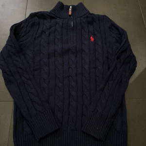 Mörkblå stickad polotröja från Polo Ralph Lauren - Säljer en mörkblå stickad polotröja från Polo Ralph Lauren med kabelstickat mönster och halv dragkedja. Tröjan har lång ärm och röd broderad logga på bröstet. Perfekt för kyliga dagar.