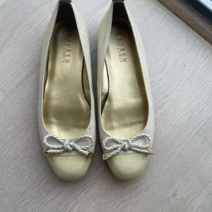 Beige ballerinaskor med rosett från Lauren Ralph Lauren - Snygga beige ballerinaskor från Lauren Ralph Lauren med lackad ljusgul tå och dekorativ rosett framtill. Skorna har rund tå och platt sula, perfekt för en stilren look.