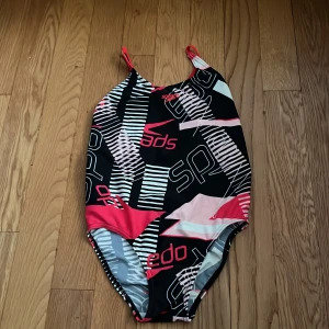 Färgglad baddräkt från Speedo - Säljer en sportig baddräkt från Speedo med svart, vit och röda mönster och tunna axelband. Perfekt för simträning eller bad. Snygg design med öppen rygg och knytning baktill.