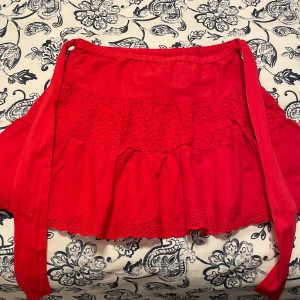 Röd kjol - Säljer en röd omlottkjol i storlek 38. Kjolen har broderade detaljer och volangkant, samt knytband i midjan. 