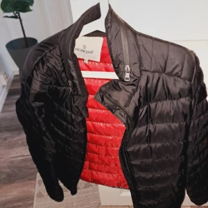 Svart dunjacka från Moncler med röd insida - Säljer en svart dunjacka från Moncler med snygg röd insida. Jackan har dragkedja framtill och är lätt vadderad, perfekt för kyliga dagar. Klassisk design med lång ärm och diskreta sömmar. Passar dig som vill ha en stilren och exklusiv look. Passar perfekt vår/höst/vinter