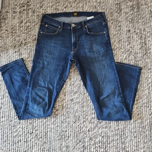 Blå jeansbyxor från Lee Luke W32 L34 - Säljer ett par klassiska blå jeans från Lee, modell Luke. Byxorna har raka ben, fem fickor och snygga kontrastsömmar. Perfekta för dig som gillar en tidlös jeansstil. Midjan är normal och passformen är straight. 
