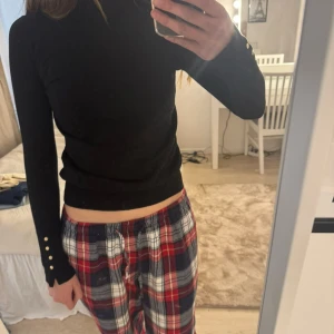 Svart polotröja med knappdetaljer - Säljer en svart polotröja med långa ärmar och snygga guldfärgade knappar vid ärmsluten. Tröjan är tight och har en klassisk, stilren look som passar till mycket. Perfekt basplagg för garderoben.