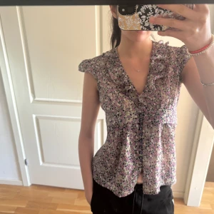 Blommig volangblus med knytband - Söt blommig blus i lila, rosa och grönt med volangdetaljer längs kragen och framtill. Blusen har knappar framtill, korta ärmar och ett knytband i midjan för en feminin siluett. Perfekt för vår och sommar!