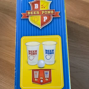 Beer Pong spelset - Säljer ett Beer Pong spelset med 14 delar. Innehåller plastmuggar i vitt med röd och blå text samt pingisbollar. Perfekt för att spela det klassiska dryckesspelet med vänner. Förpackningen är blå med färgglada detaljer och instruktioner på sidan.