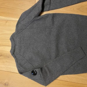 Grå stickad tröja från C.P. Company - Säljer en stilren grå stickad tröja från C.P. Company med deras ikoniska linsdetalj på ärmen. Tröjan har rund halsringning och långa ärmar med ribbade muddar och nederkant. ,XS/XXS.