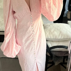 Ljusrosa adoore klänning - Superfin ljusrosa långklänning med djup v-ringning, puffiga långärmar och dekorativa knappar framtill. Klänningen har ett subtilt blommönster i tyget och en slits framtill för extra detalj. Helt ny! Precis fått hem, lapp kvar!