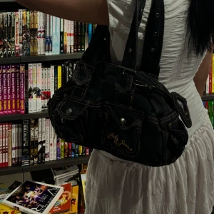 Svart handväska från Baby Jeans med jeansdetaljer - Cool svart handväska från Baby Jeans i jeanstyg med kontrastsömmar och flera fickor framtill. Väskan har dubbla handtag med spännen och en hjärtformad spegel som accessoar. Perfekt för dig som gillar en unik och trendig look.