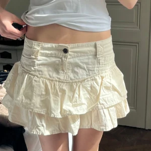 Vit/ beige volangkjol från Bershka - Supersöt vit kortkjol från Bershka med dubbla volanger och knapp framtill. Kjolen har klassiska jeansdetaljer som fickor.