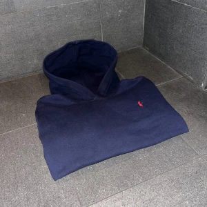Mörkblå hoodie från Polo Ralph Lauren - Liten i storleken passar någon som kanske är runt 155-165. Säljer en mörkblå hoodie från Polo Ralph Lauren med klassisk röd broderad logga på bröstet. Tröjan har huva och magficka, samt ribbade muddar vid ärmslut och nederkant. Perfekt för en avslappnad och stilren look. Tröjan är i bra skick och priset kan alltid sänkas