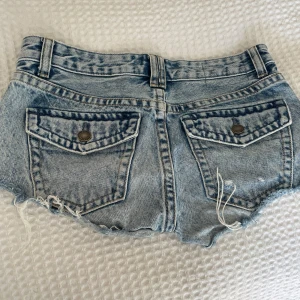 Blå jeansshorts med fransar - Säljer ett par blå jeansshorts med råa fransade kanter och bakfickor med knapp. Shortsen har bälteshällor och klassisk femficksdesign. Perfekta för varma dagar och har en avslappnad, trendig look.