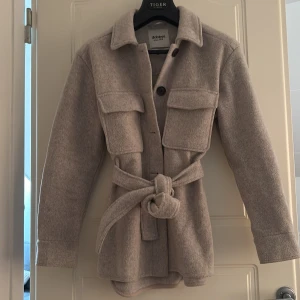 Beige overshirt med bälte från dobber  - Snygg beige overshirt från Dobber med bälte i midjan och stora bröstfickor. Jackan har lång ärm, knappar framtill och är tillverkad i ett mjukt material som känns lite ulligt. Perfekt för lager-på-lager-stil under höst och vår.