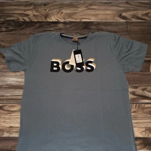 Grå t-shirt med BOSS-tryck - Snygg grå t-shirt från BOSS med stort svart och vitt logotyptryck på bröstet. Klassisk rund halsringning och korta ärmar. Perfekt för en stilren och avslappnad look.