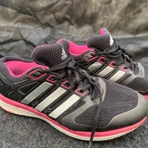 Svarta Adidas löparskor med rosa detaljer - Säljer ett par svarta Adidas sneakers med rosa och vita detaljer. Skorna har klassiska tre ränder på sidan, svart mesh-överdel och rosa insida. Perfekta för träning eller vardagsstil. Skön passform med snörning och platt sula.