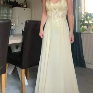 Gul långklänning med spetsdetaljer - Gul långklänning från Mascara med spets upptill och flowy kjol med slitts. Klänningen är ärmlös och har markerad midja. Klänningen är insydd till en 32/34, det går att sy ut klänningen igen då inget tyg klipptes av. Jag är 162 och hade klackskor vid användning och fötterna syntes inte. Klänningen har fläckar ner till men som inte syns om du inte lyfter på klänningen, på baksidan av klänningen har det blivit indragningar i tyget som visas på bild 3/4 (kan skicka mer bilder vid efterfrågan).