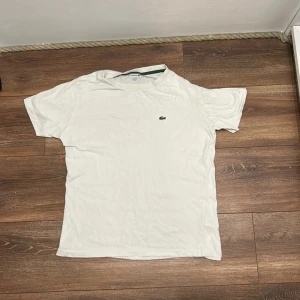 Vit t-shirt från Lacoste - Vit t-shirt från Lacoste med klassisk grön krokodil-logga broderad på bröstet. T-shirten har rund hals och korta ärmar, perfekt för en clean och stilren look.