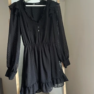 Svart volangklänning från Zara - Säljer en svart kortklänning från Zara med volangdetaljer vid ärmslut och nederkant. Klänningen har långärmade transparenta ärmar, knappar framtill och diskret struktur i tyget. St. S. Använd en gång 